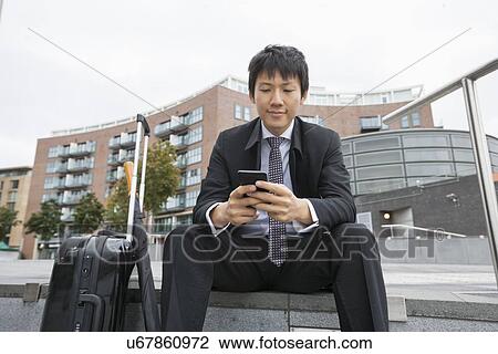 Asian businessman with luggage text messaging through cell phone against buildings View Large Photo Image Stock Image - Asian businessman with luggage text messaging through cell phone against buildings. Fotosearch