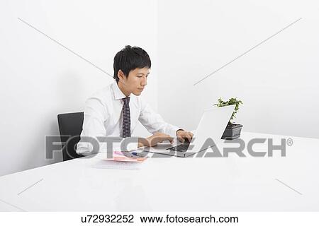 Stock Image - Asian mid adult businessman using laptop at desk in office. Fotosearch
