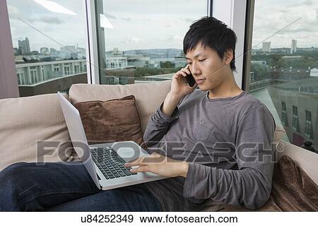 Stock Photo - Asian mid adult man on call while using laptop in living room. Fotosearch