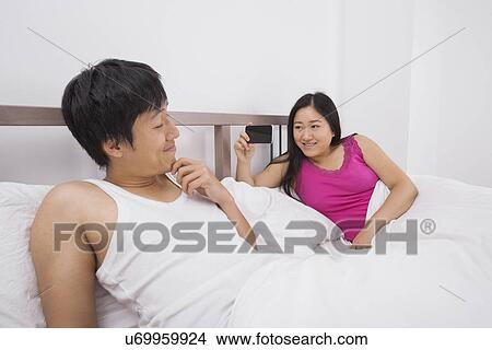 Picture - Asian woman taking a picture of man with cell phone in bed. Fotosearch