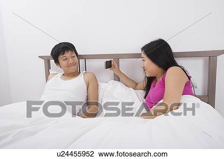 Stock Image - Asian woman taking a picture of man with cell phone in bed. Fotosearch