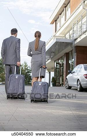 Stock Image - Back view of businesspeople walking with luggage outside hotel. Fotosearch