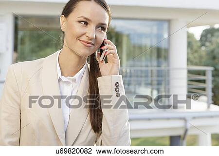 Stock Image - Beautiful businesswoman answering cell phone against office building. Fotosearch