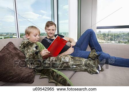 Stock Photography - Boy dressed in dinosaur costume reading picture book with father on sofa bed at home. Fotosearch