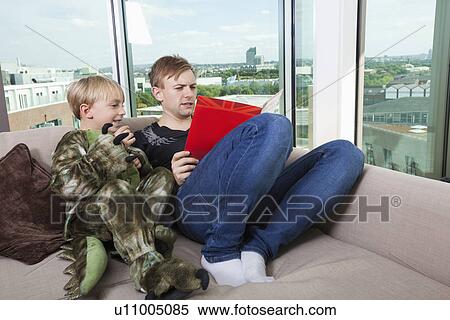 Stock Photography - Boy dressed in dinosaur costume sitting with father reading story book on sofa bed at home. Fotosearch