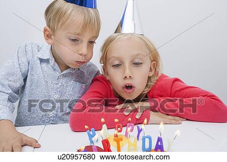Stock Image - Brother and sister blowing birthday candles at table in house. Fotosearch
