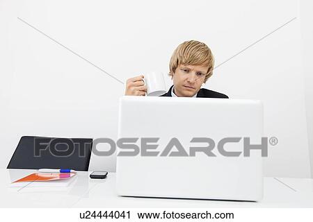 Stock Image - Businessman having coffee while using laptop in office. Fotosearch