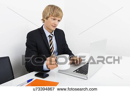 Businessman holding coffee cup while using laptop at desk in office View Large Photo Image Stock Photo - Businessman holding coffee cup while using laptop at desk in office. Fotosearch