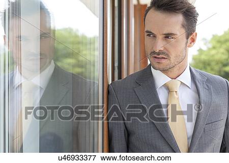 Stock Photo - Businessman looking away while leaning on glass door. Fotosearch