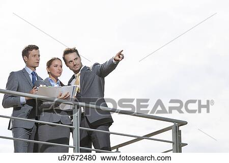 Businessman showing something to coworkers on terrace against sky View Large Photo Image Stock Image - Businessman showing something to coworkers on terrace against sky. Fotosearch