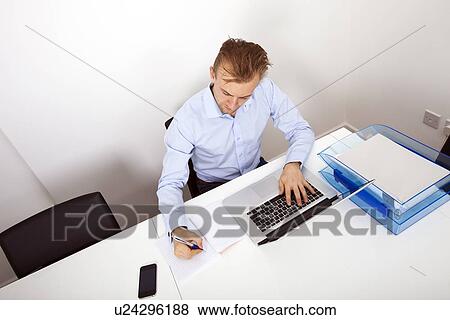 Stock Photo - Businessman taking notes while using laptop in office. Fotosearch