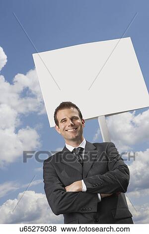 Businessman with arms crossed standing by blank sign against cloudy sky View Large Photo Image Stock Photo - Businessman with arms crossed standing by blank sign against cloudy sky. Fotosearch