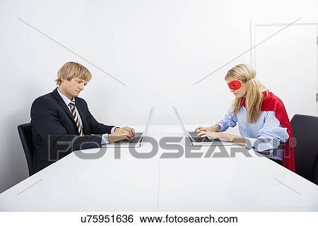 Stock Photograph - Businessman with coworker in superhero costume using laptops at office. Fotosearch