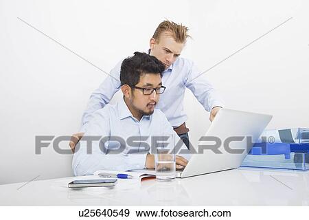 Businessmen using laptop together at desk in office View Large Photo Image Stock Photo - Businessmen using laptop together at desk in office. Fotosearch