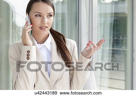 Stock Photo - Businesswoman gesturing while answering cell phone against glass door. Fotosearch