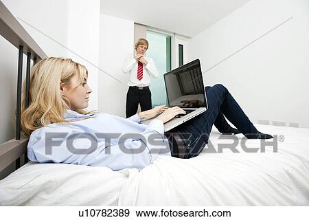 Stock Photo - Businesswoman using laptop in bed with businessman adjusting tie at hotel. Fotosearch