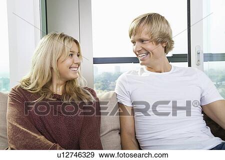 Cheerful couple sitting on sofa and looking at each other at home View Large Photo Image Stock Photo - Cheerful couple sitting on sofa and looking at each other at home. Fotosearch