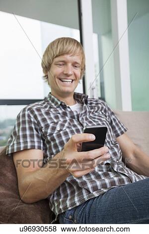 Cheerful mid-adult man text messaging on sofa at home View Large Photo Image Stock Photo - Cheerful mid-adult man text messaging on sofa at home. Fotosearch