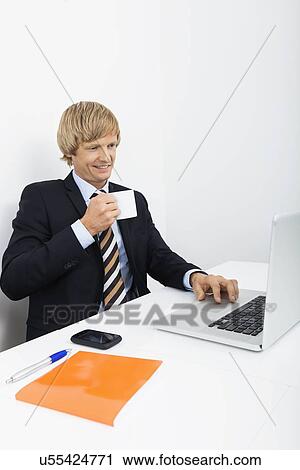 Confident businessman having coffee white using laptop in office View Large Photo Image Stock Image - Confident businessman having coffee white using laptop in office. Fotosearch