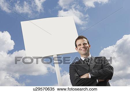 Confident businessman with arms crossed standing by blank sign against cloudy sky View Large Photo Image Stock Photography - Confident businessman with arms crossed standing by blank sign against cloudy sky. Fotosearch