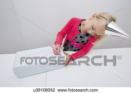 Stock Image - Cute girl writing on birthday gift at table in house. Fotosearch