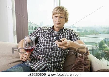 Displeased mid-adult man with wine glass watching television on sofa at home View Large Photo Image Stock Photo - Displeased mid-adult man with wine glass watching television on sofa at home. Fotosearch