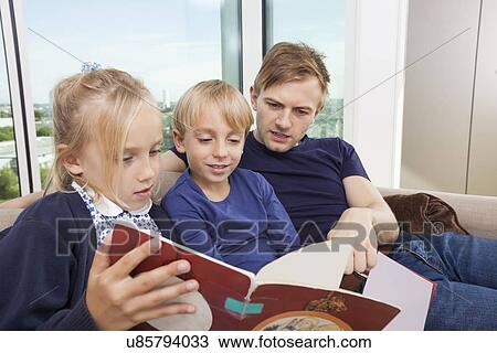 Stock Image - Father and children reading storybook on sofa at home. Fotosearch