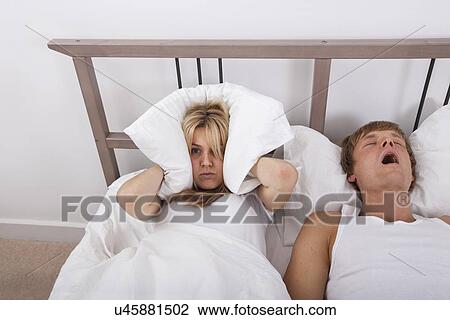 Stock Image - Frustrated woman covering ears with pillow while man snoring in bed. Fotosearch