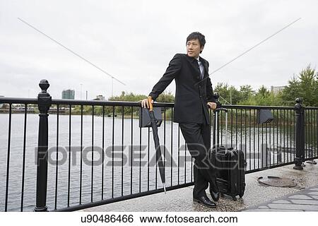 Stock Photograph - Full length of businessman with luggage leaning on railing along river. Fotosearch