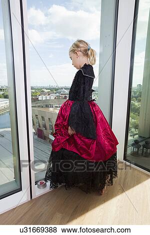Stock Photo - Full length of girl in vampire costume looking out through window at home. Fotosearch