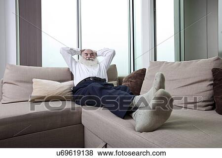 Stock Photo - Full length of senior man reclining on sofa with hands behind head at home. Fotosearch