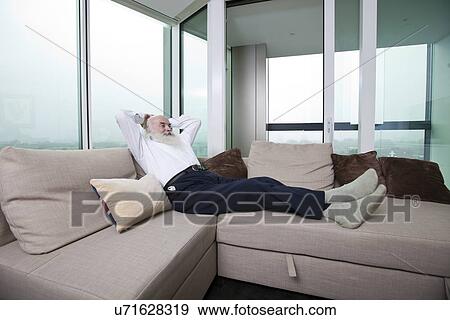 Stock Photo - Full length of senior man relaxing on sofa with hands behind head at home. Fotosearch