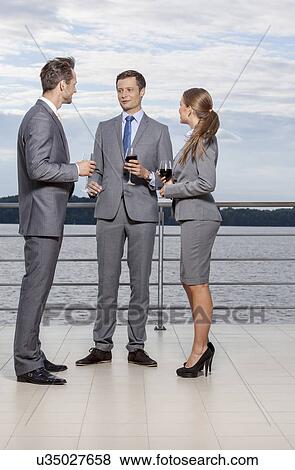 Full length of young businesspeople holding wineglasses while communicating on terrace View Large Photo Image Stock Photo - Full length of young businesspeople holding wineglasses while communicating on terrace. Fotosearch