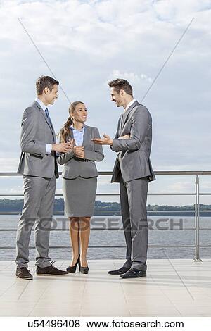 Stock Photo - Full length of young businesspeople communicating on terrace against sky. Fotosearch