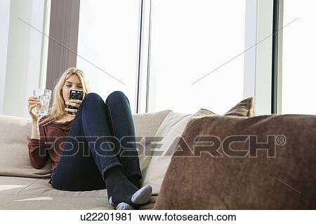Stock Image - Full length of young woman text messaging at home. Fotosearch