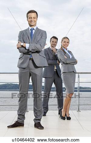 Stock Image - Full length portrait of confident businessman standing with coworkers on terrace against sky. Fotosearch