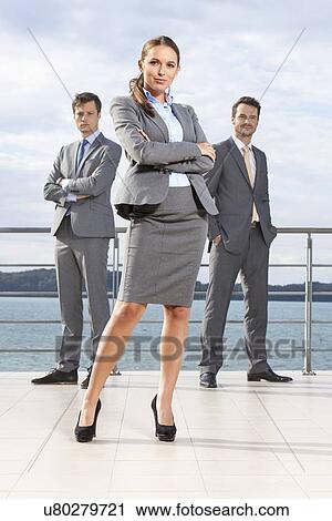 Stock Image - Full length portrait of confident businesswoman standing with coworkers on terrace against sky. Fotosearch