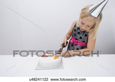 Stock Image - Girl cutting birthday cake at table in house. Fotosearch