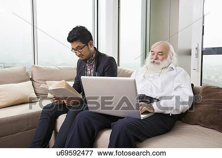 Stock Photo - Grandfather using laptop while grandson reading book on sofa at home. Fotosearch