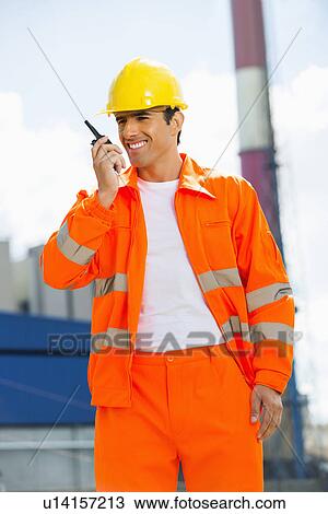Stock Image - Happy architect wearing reflective workwear communicating on walkie-talkie at site. Fotosearch