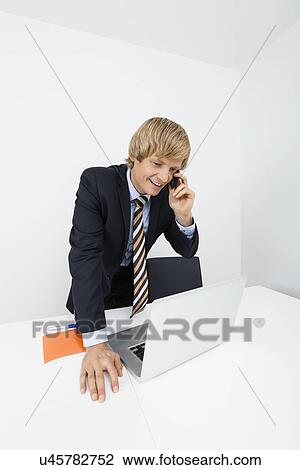 Stock Image - Happy businessman using cell phone while looking at laptop in office. Fotosearch