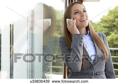 Stock Photo - Happy businesswoman answering cell phone by glass door. Fotosearch