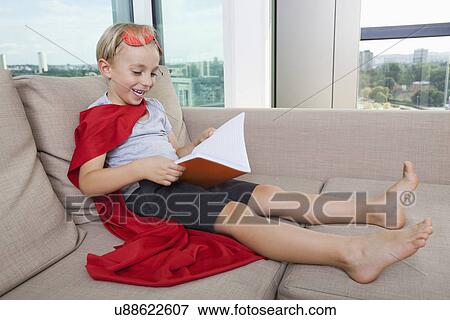 Stock Photo - Happy little boy in superhero costume reading book on sofa at home. Fotosearch