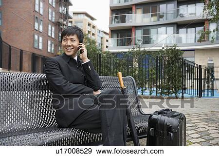 Stock Photo - Happy mid adult businessman with luggage using cell phone on bench. Fotosearch