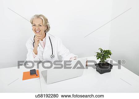 Stock Image - Happy senior female doctor with laptop at desk in clinic. Fotosearch