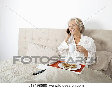 Stock Image - Happy senior woman answering cell phone while having breakfast in bed. Fotosearch