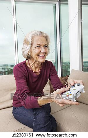 Stock Image - Happy senior woman playing video game on sofa at home. Fotosearch