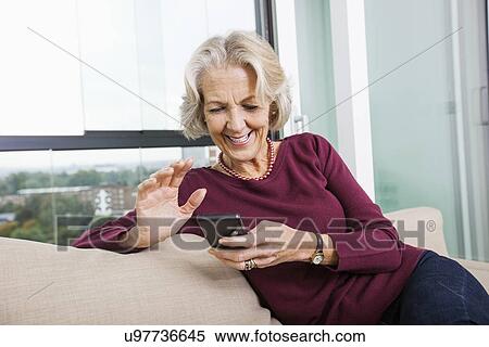 Happy senior woman text messaging through smart phone on sofa at home View Large Photo Image Stock Photography - Happy senior woman text messaging through smart phone on sofa at home. Fotosearch