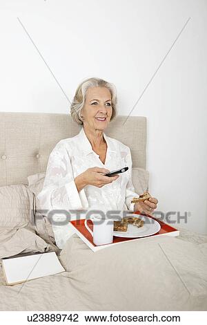Stock Image - Happy senior woman watching TV while having breakfast in bed. Fotosearch