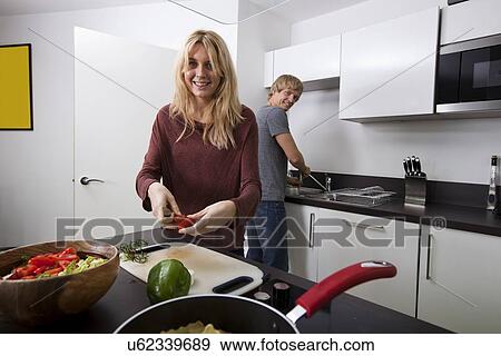 Stock Photo - Happy woman preparing food with man standing in background at kitchen. Fotosearch
